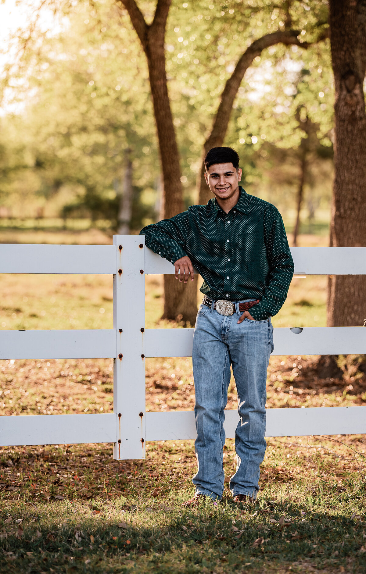 A senior boy wearing a green collared shirt leans against a white fence at Jack Brooks Park.