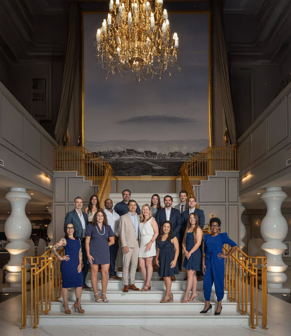Professional group portrait by St. Augustine headshot photographer featuring nine people in coordinated blue and white attire posed on an elegant curved staircase with ornate railings and classical architecture