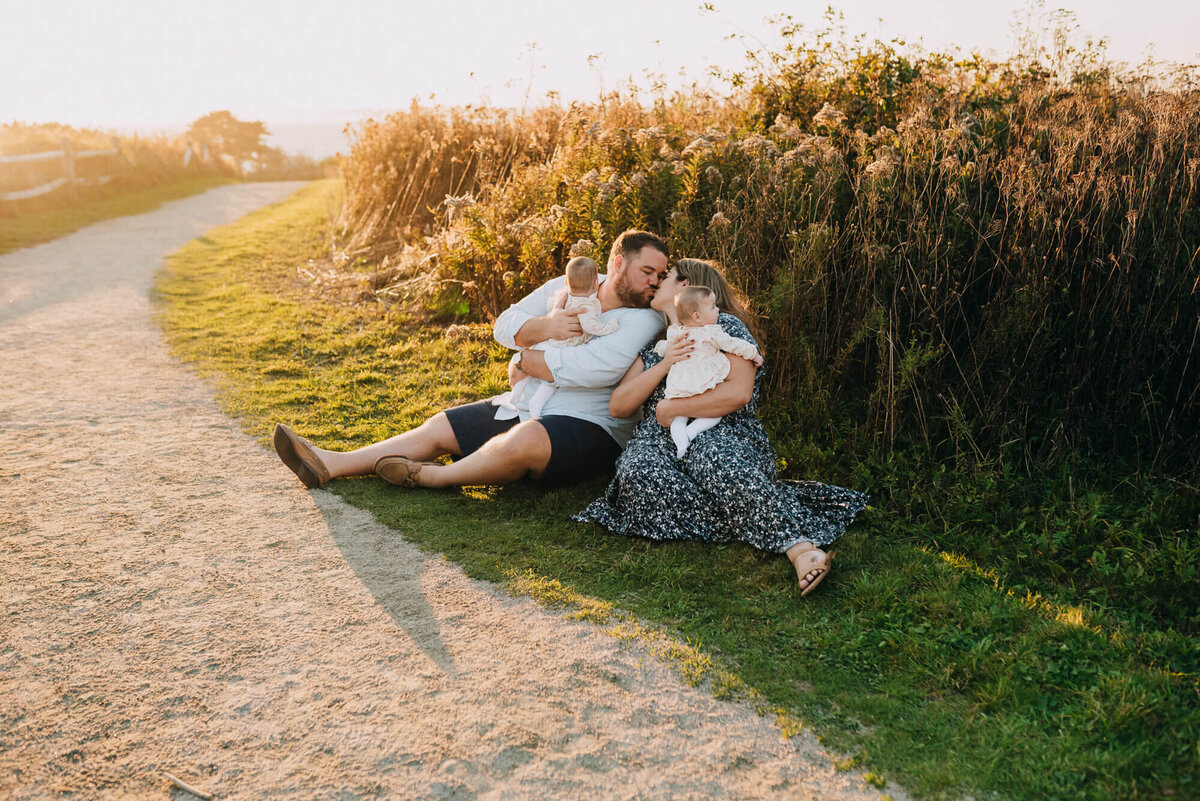 A mother and father sit on the ground near a path close to the beach at sunset