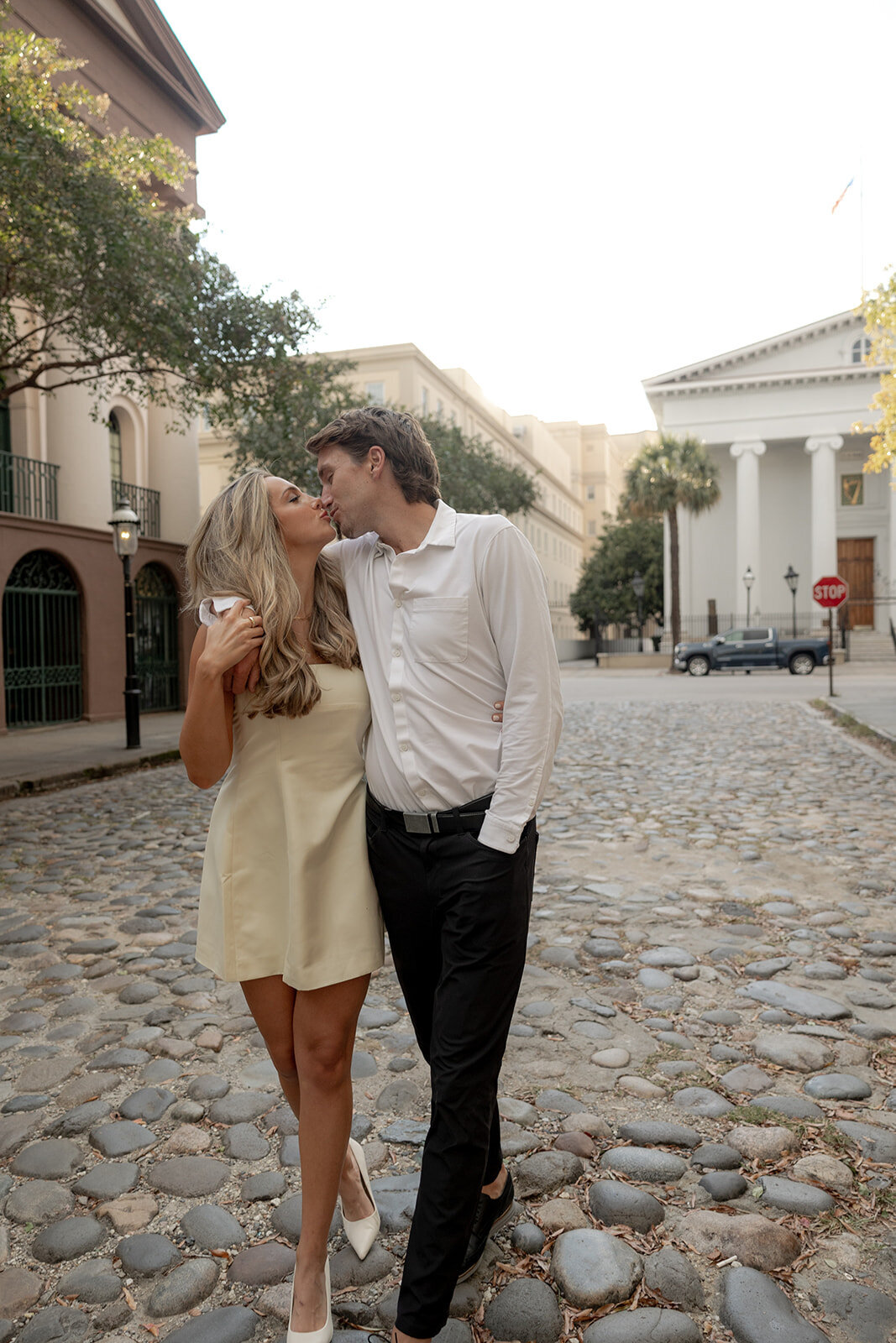 Couple kissing while walking on Charleston cobblestone street. Man in black pants and white shirt. Woman with champagne dress for engagement session.