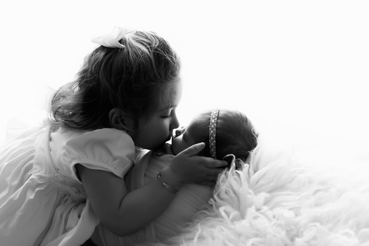 Toddler leaning over her newborn sister tenderly kissing her on a white background.