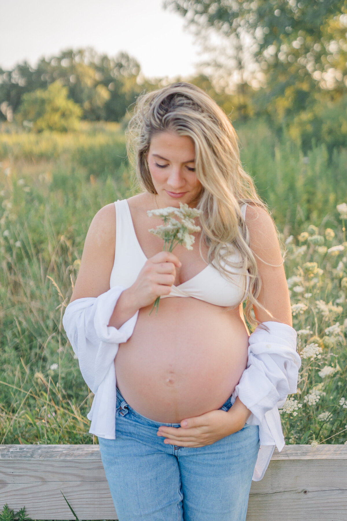 a mother stands in a beautiful field with her pregnant belly exposed while smelling a flower