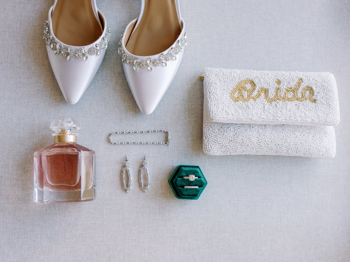 A flat lay of the brides personal details including her wedding shoes, rings, perfume, jewelry, and purse