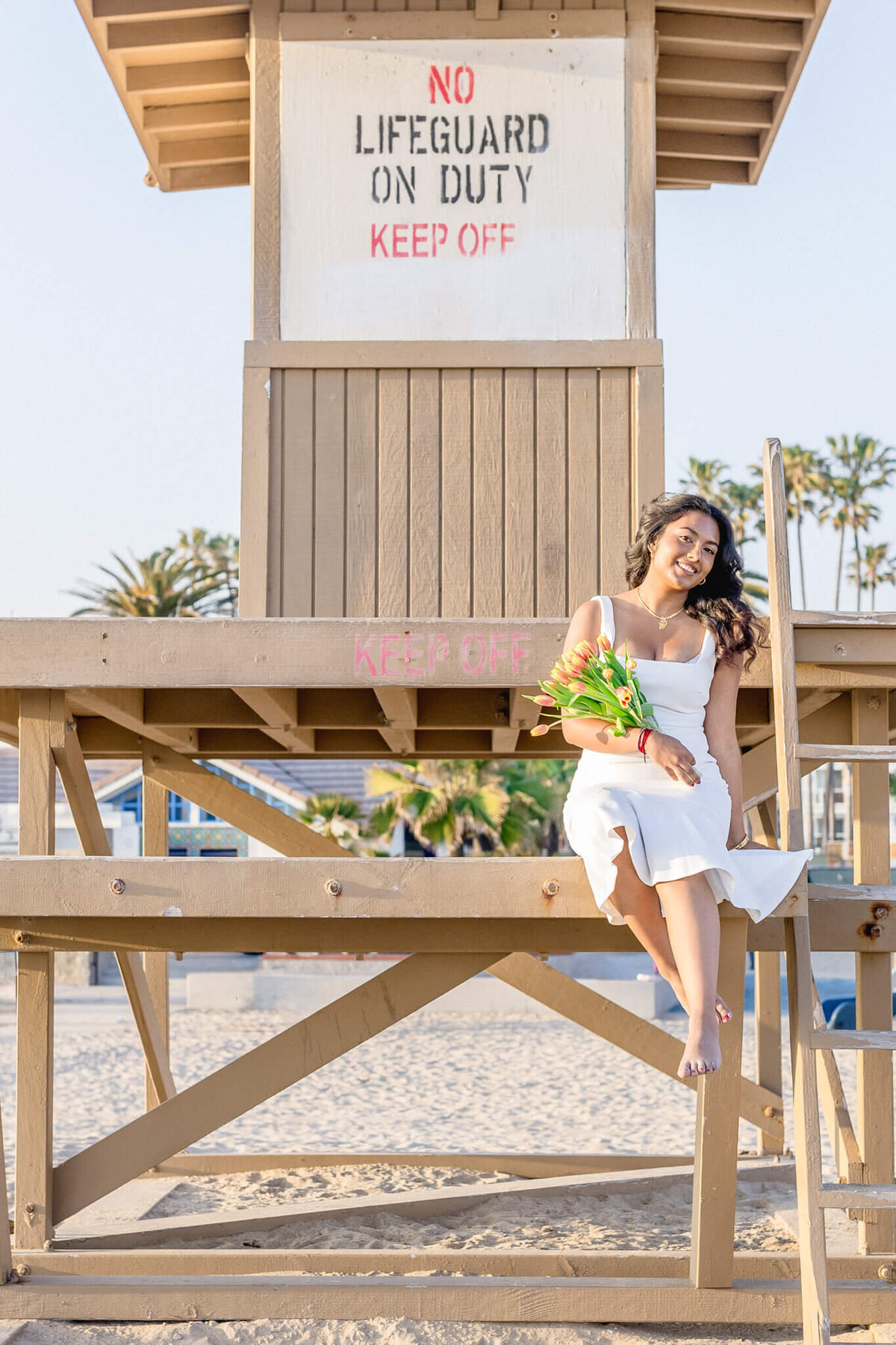 A woman in a white dress sits on a beach lifeguard tower holding a bouquet of yellow flowers. The tower has No lifeguard on duty, keep off signs. The background features palm trees and a clear sky.