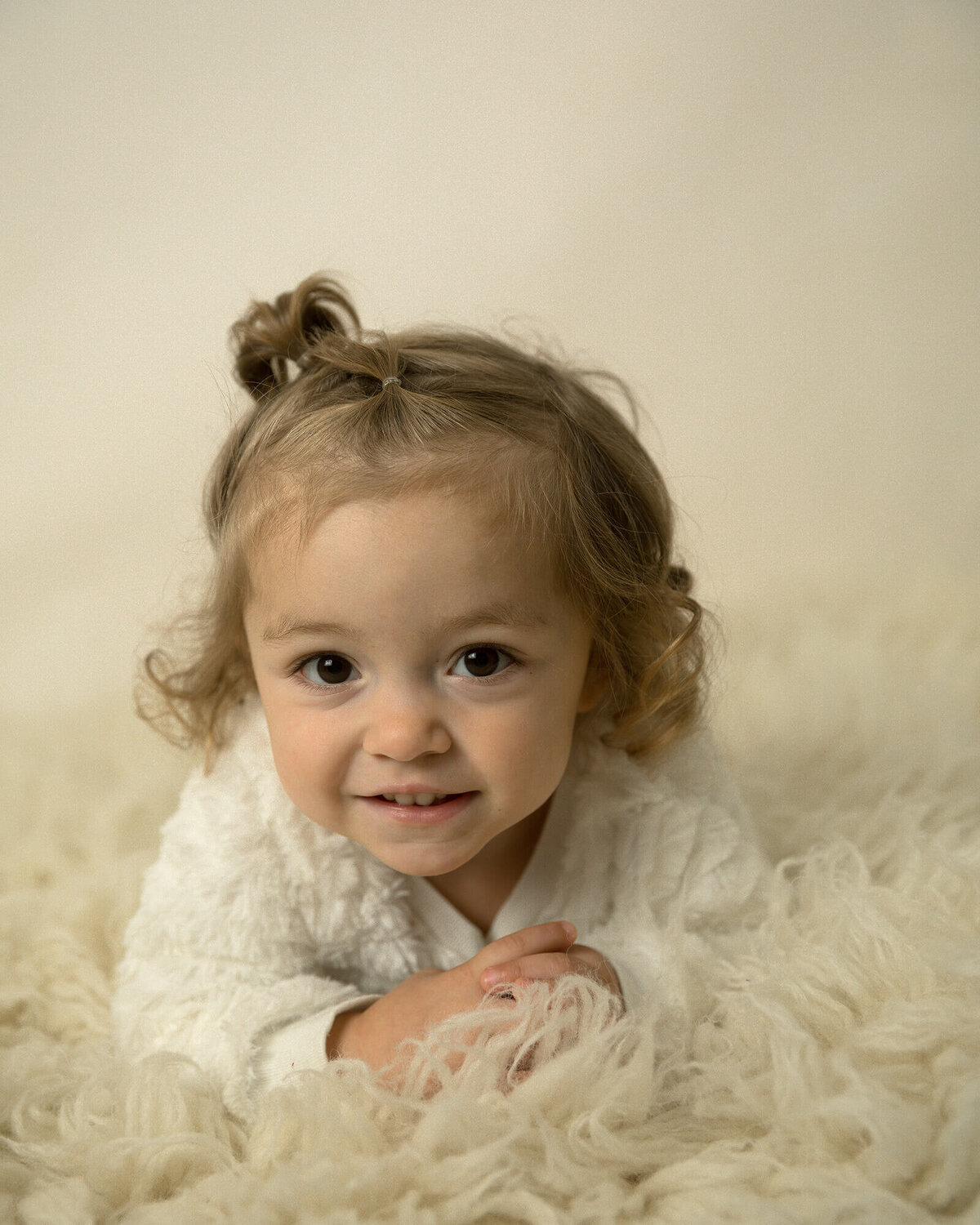 Little girl with curly brown hair laying on a creme colored rug.
