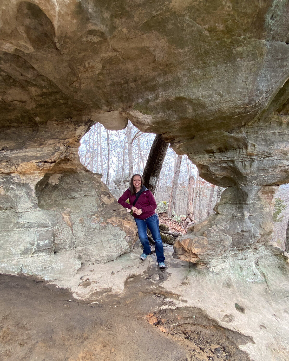 woman sitting on a rock formation smiling at the camera, wearing a jacket, pants, and hiking boots