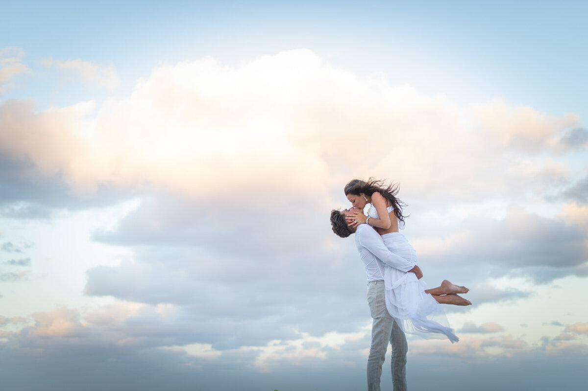 A couple shares a joyful moment as one lifts the other against a vibrant sunset sky.