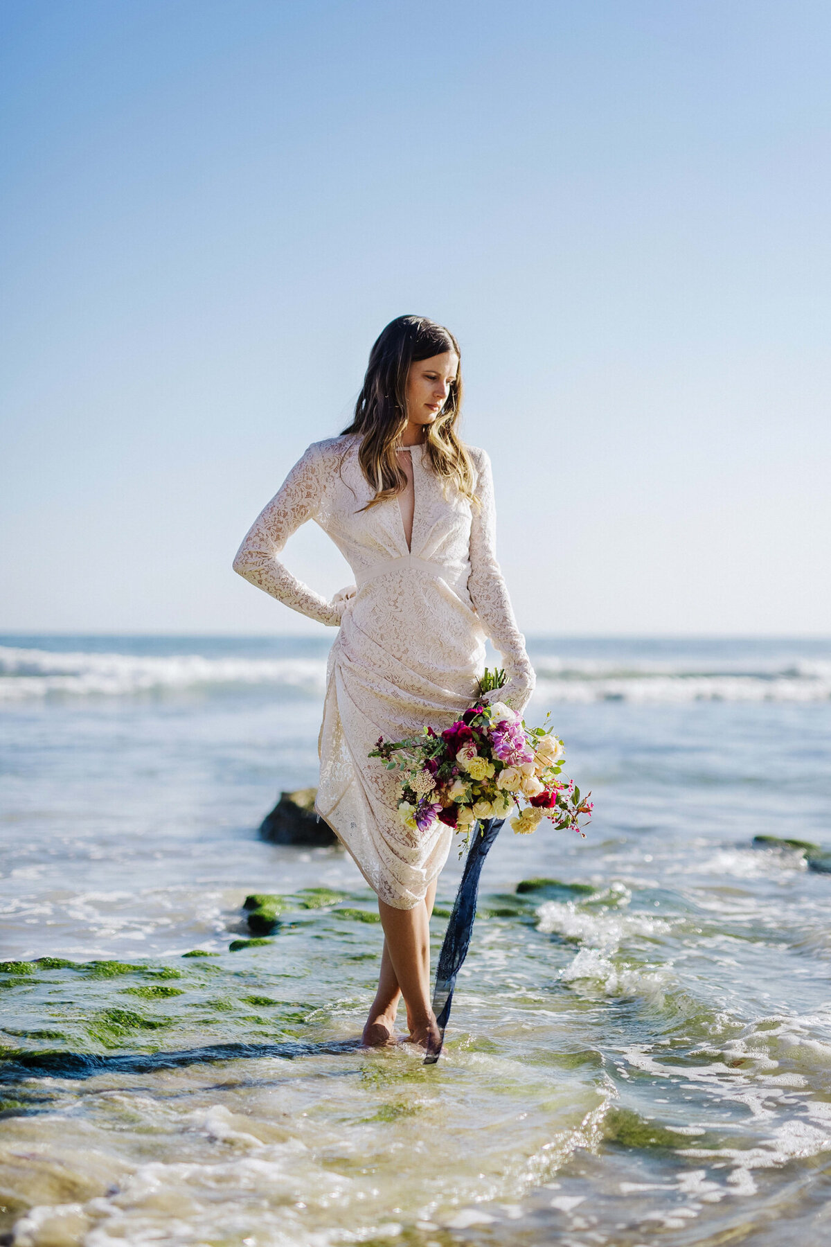 Local Calgary wedding inspiration, bride standing in water, wearing a vintage long-sleeved lace gown, holding a beautiful bouquet of yellow and purple flowers held together with a blue satin ribbon, captured by Christy D. Swanberg Photography, editorial elopement and wedding photographer in Calgary, Alberta, featured on the Bronte Bride Vendor Guide.