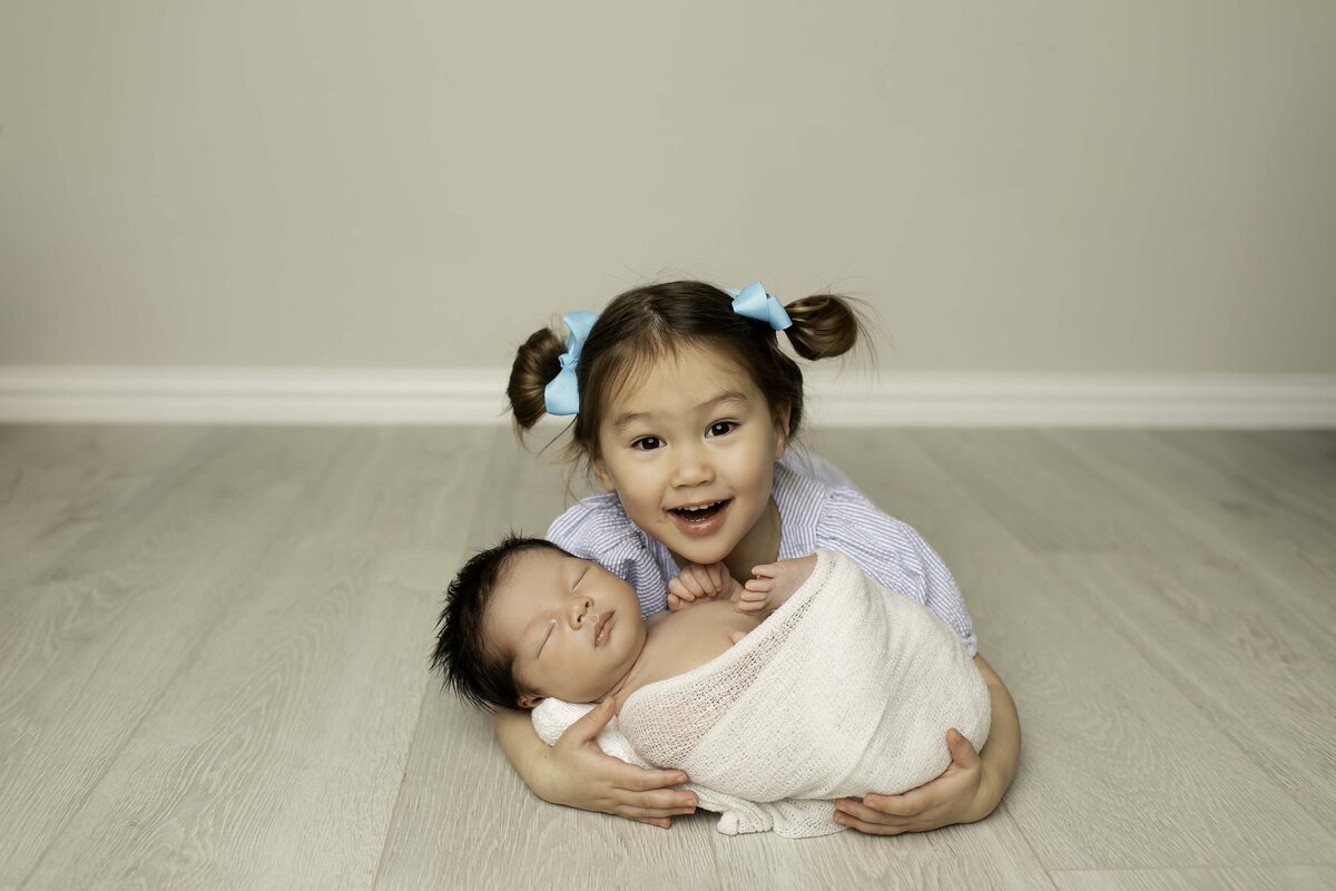 Adorable big sister with playful pigtails lovingly cradling her tiny newborn brother in her arms, both sharing a sweet sibling moment.