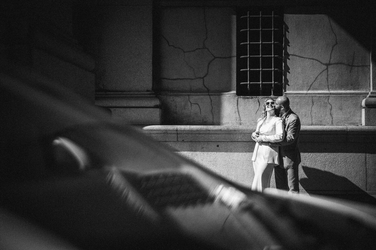 Black and white photo of a couple embracing in a shaft of sunlight against a textured stone wall near the NYC Marriage Bureau, framed by cars in motion—an expressive image by an NYC elopement photographer highlighting urban contrast and emotion.