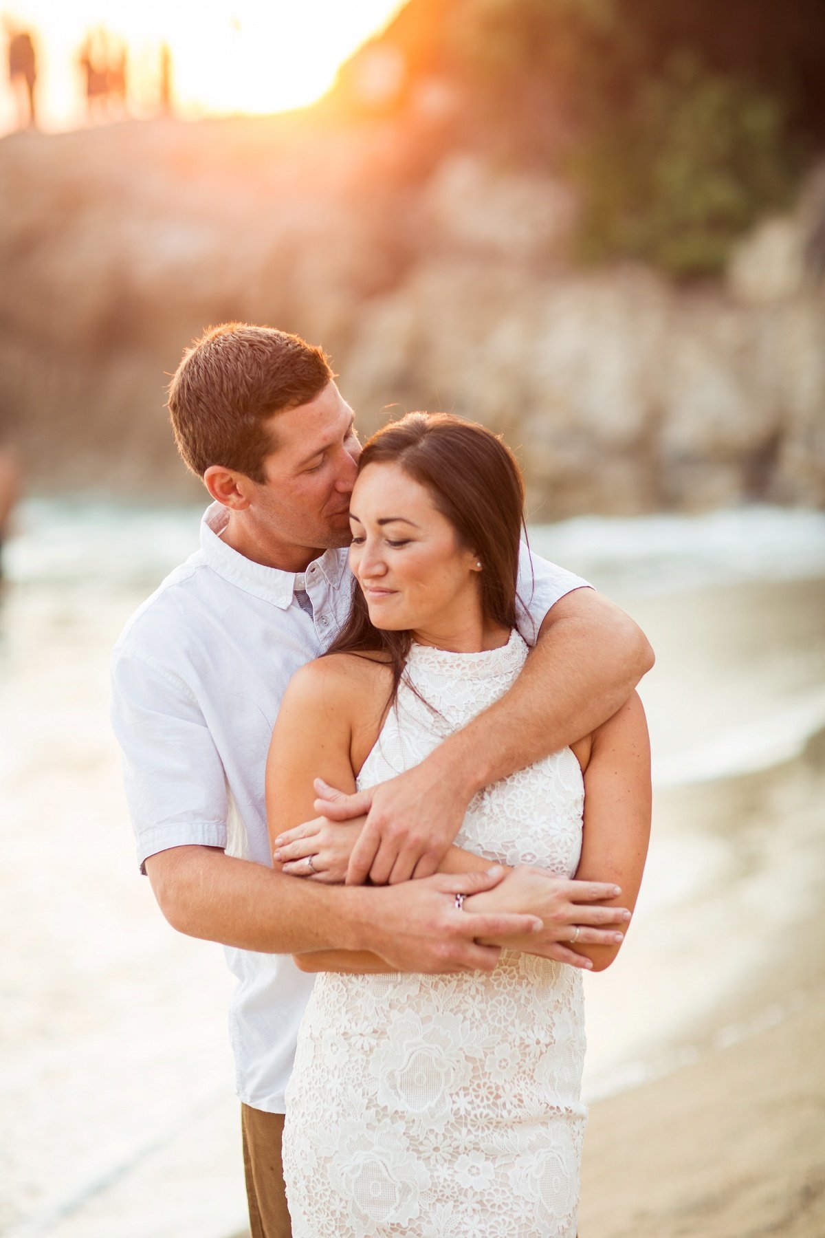 Groom to be fully embraces his Bride and kisses her head while walking along the beach