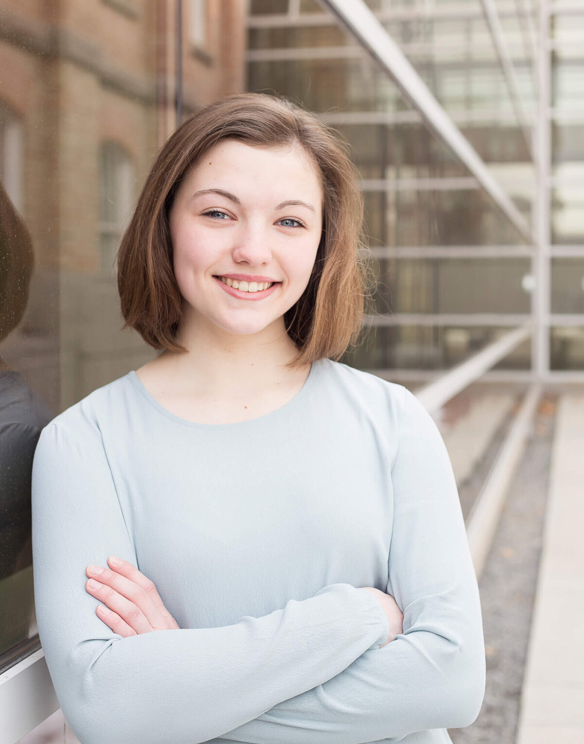 brown haired girl smiling with arms folded in front of a wall of windows