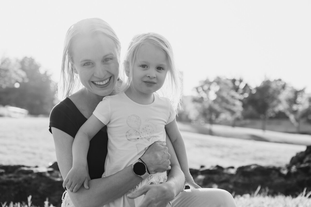 A woman laughing and holding a young girl in a park, both smiling brightly in the sunlight.