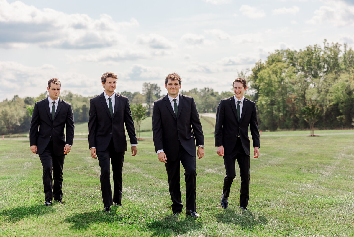 Four men in black suits and ties walk confidently across a grassy field under a partly cloudy sky, with trees in the background. They appear relaxed, perhaps discussing rustic wedding venue packages for an upcoming celebration, their camaraderie evident in their expressions.