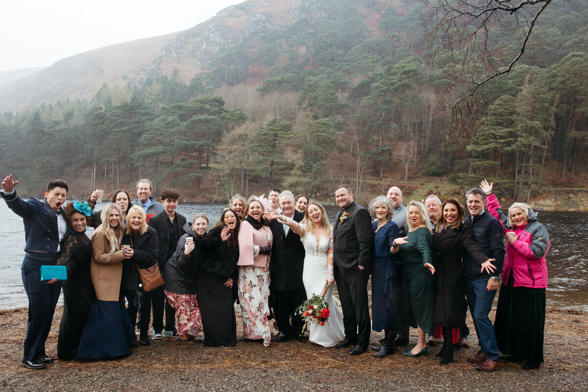 A group of wedding guests posing with the bride and groom by a lake, with hills in the background