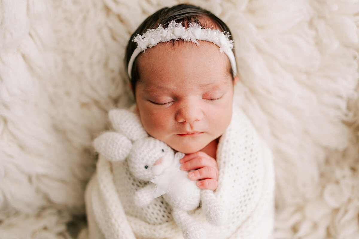 portrait of a newborn girl holding white bunny. She is sleeping on white fuzzy background and has white headband and black hair