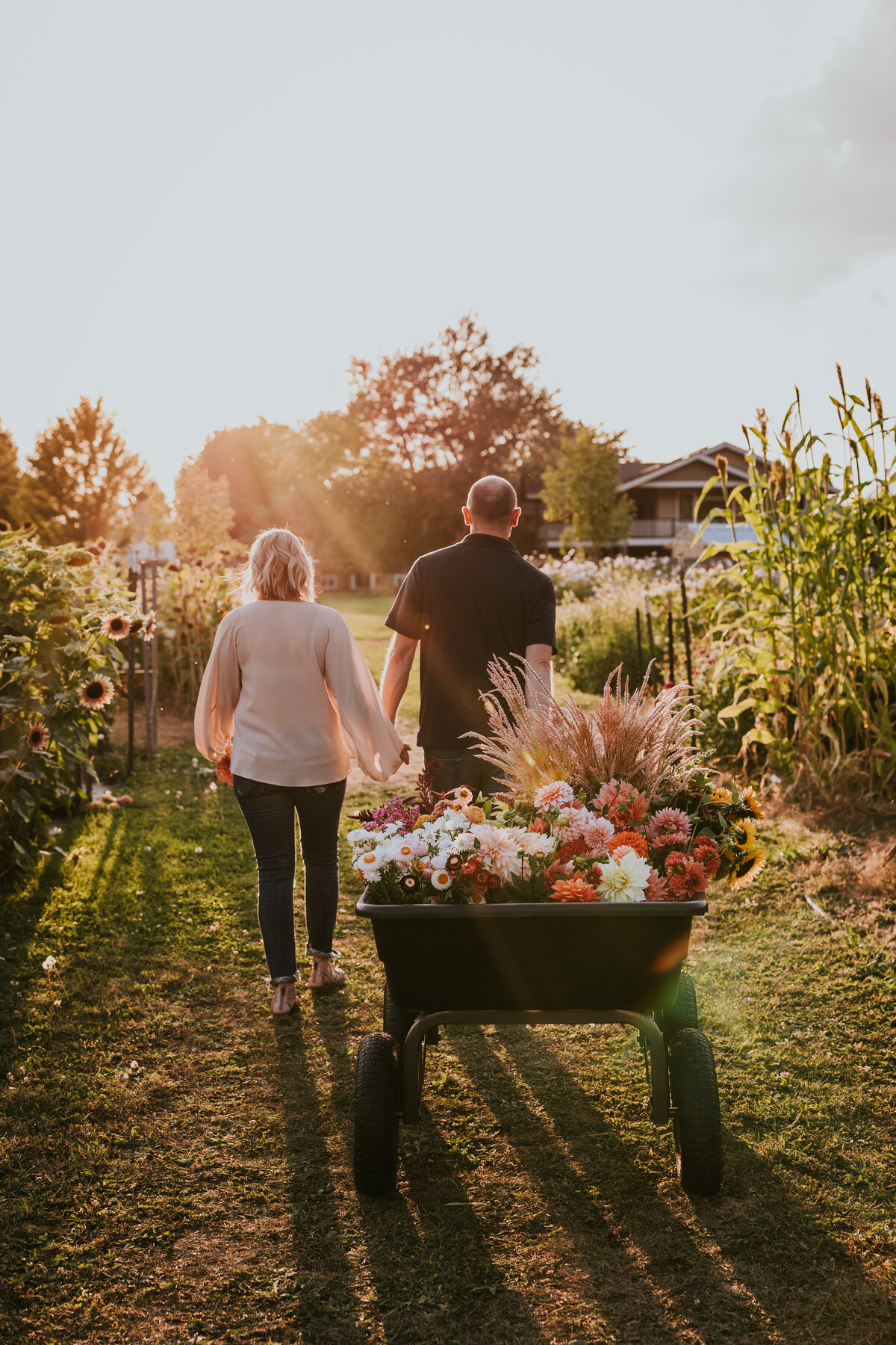 Couple holds hands while walking through garden with a wagon full of fresh flowers is in tow.