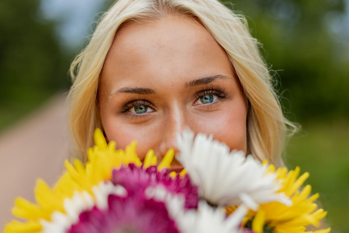 A senior girl playfully holds colorful flowers near her face, showcasing joy, bold colors, and a creative composition in this unique portrait.