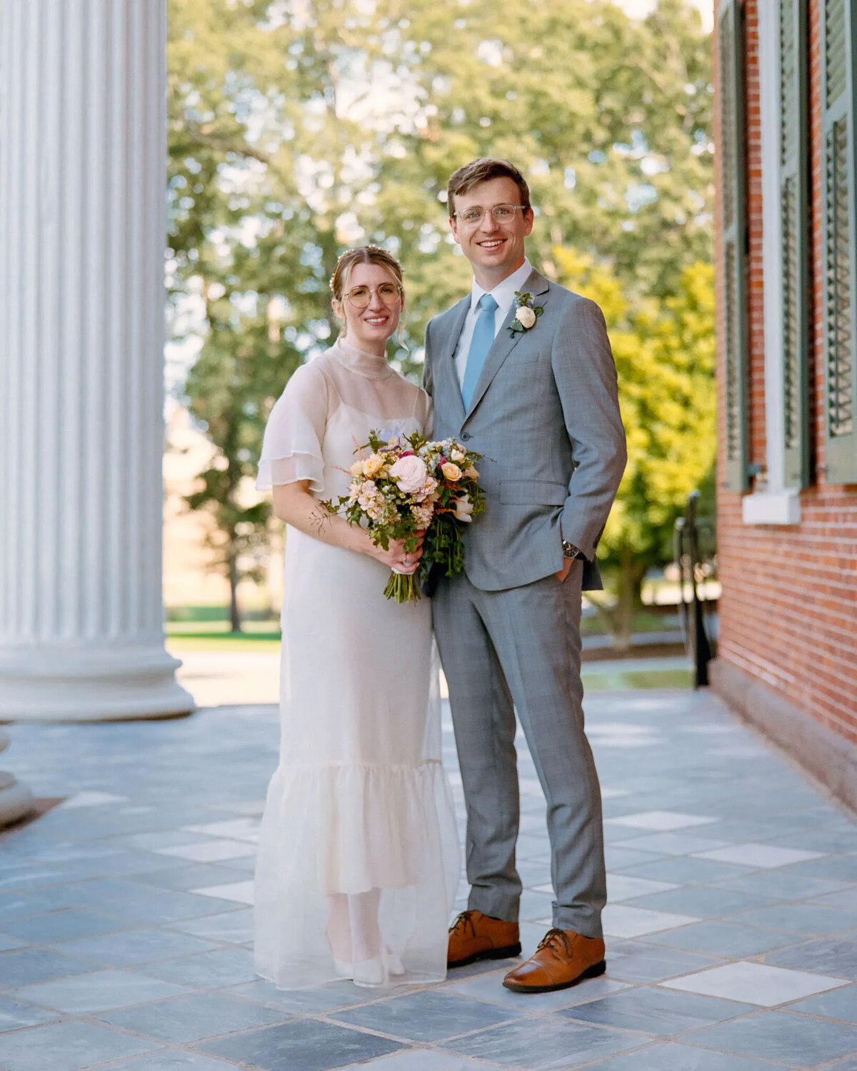 A couple stands together smiling outdoors; the woman wears a white dress and holds a bouquet, while the man wears a light gray suit and blue tie. Captured by an NJ wedding photographer, they stand on a tiled walkway beside columns and a brick building.