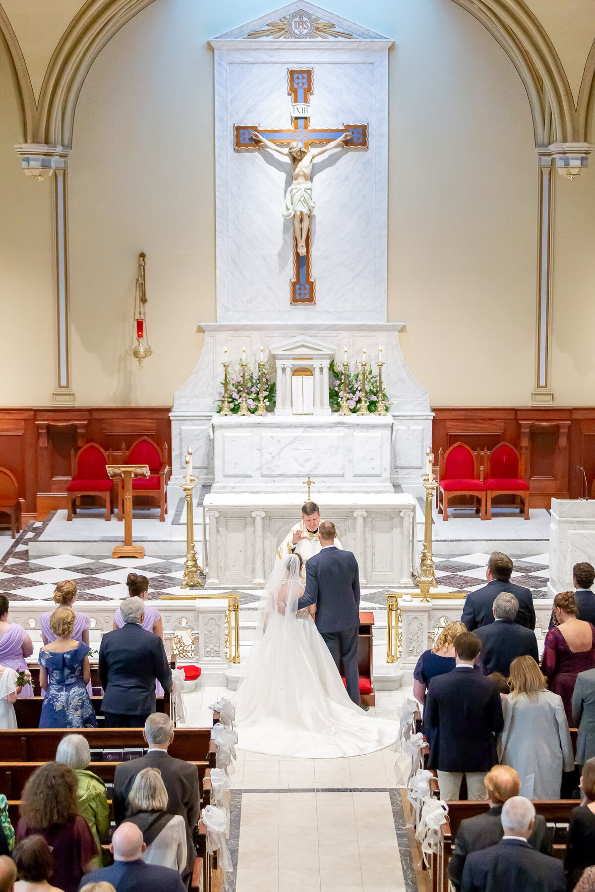 A couple stands at the alter on their wedding day inside of a church. The stands are filled with family and friends.