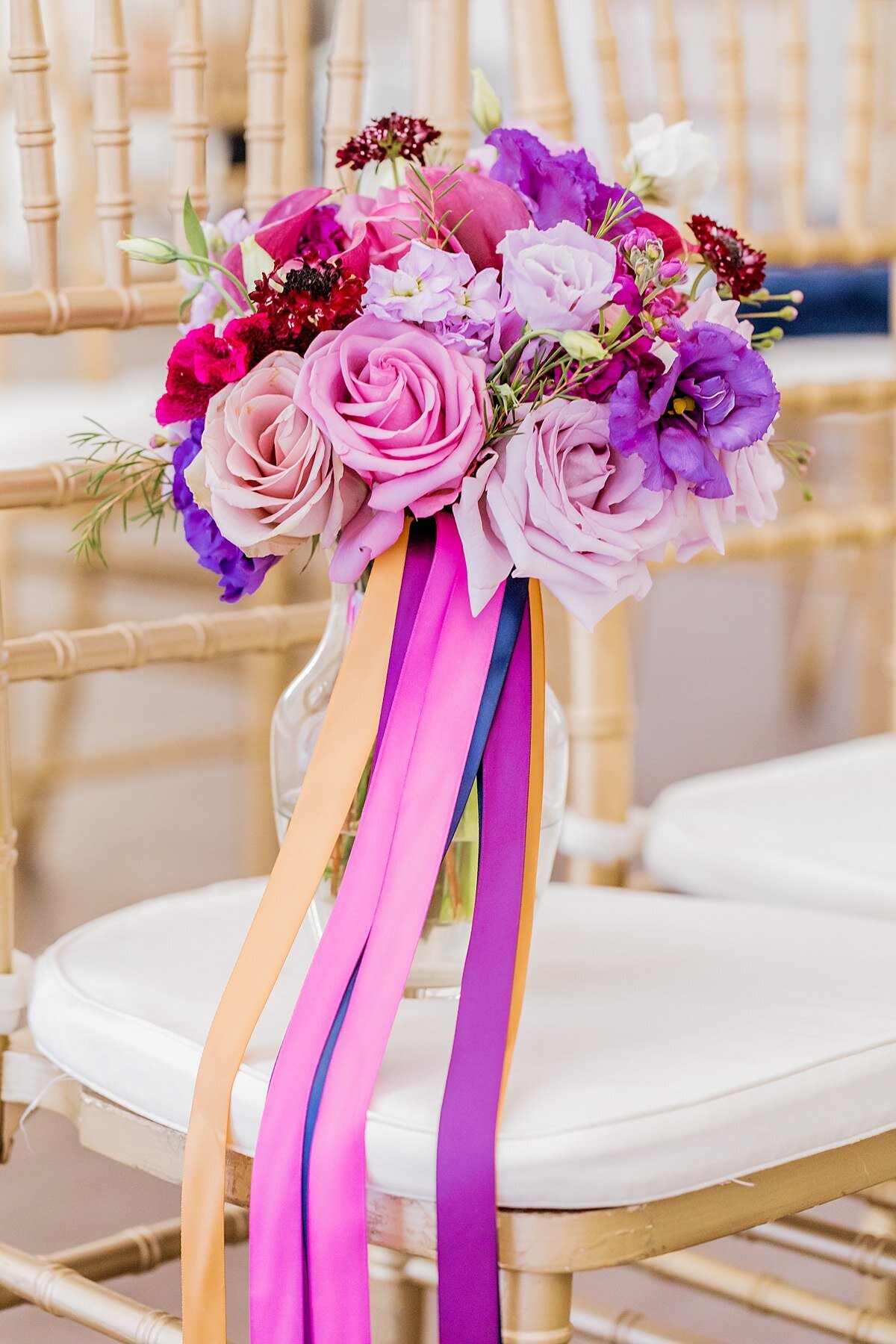 A bridal bouquet of peach roses, pink roses, magenta flowers, purple flowers and long streaming ribbons in pink, purple, navy and gold sit in a glass vase on a gold chivari chair with a white cushion.