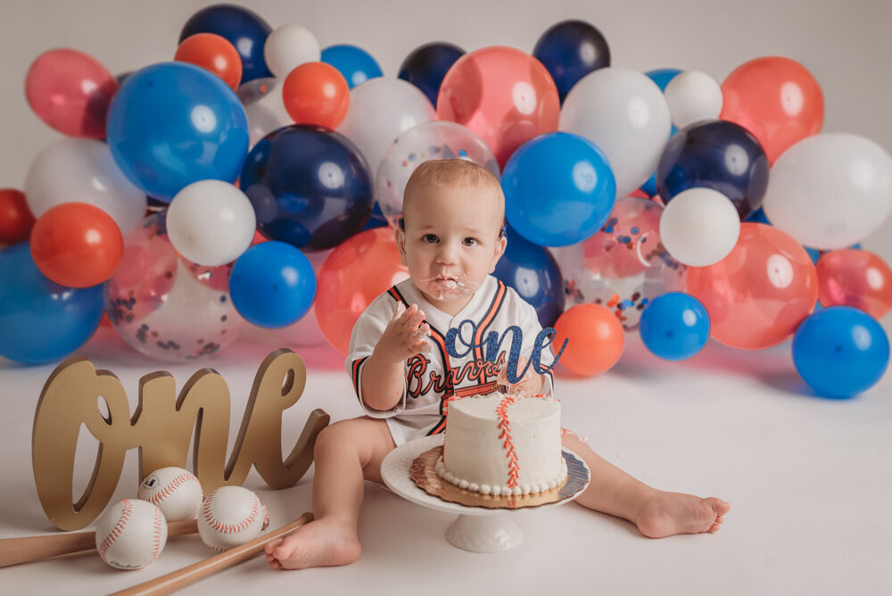 Photo shoot with Atlanta, Georgia cake smash photographer with one year old baby boy sitting on floor with an Atlanta Braves jersey on, baseballs, bats and balloons around him. He is sitting on the floor with a birthday cake in front of him and he's earing the cake.