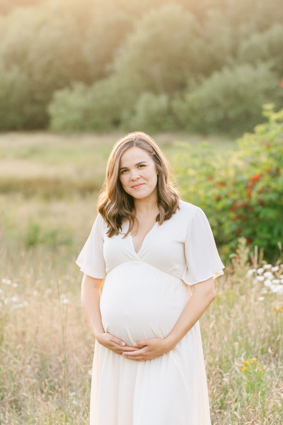 Woman with baby bump standing in a field of golden grasses wearing a cream colored dress. She is holding her bump on the bottom. She has shoulder-length brown hair and has a small grin on her face looking at the camera.