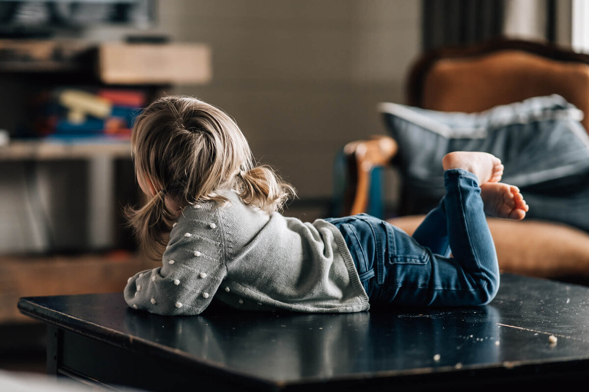 A photograph shot from behind of a little girl with pigtails lying on her tummy on the coffee table in her living room, watching TV.