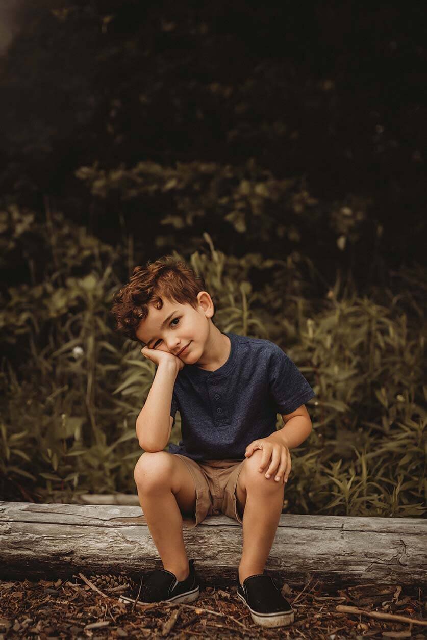 A little boy sitting on a log with one hand on his knee and the other hand is smushed against his cheek with his elbow on his knee