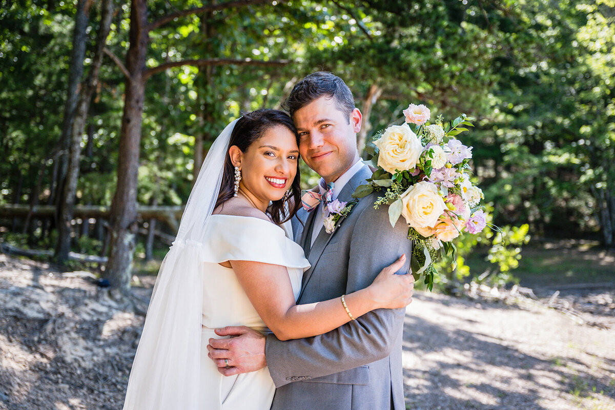 A newlywed couple hug one another and smile under a canopy of trees at Carvin’s Cove in Roanoke, Virginia. The bride’s bouquet is held against the groom’s back.