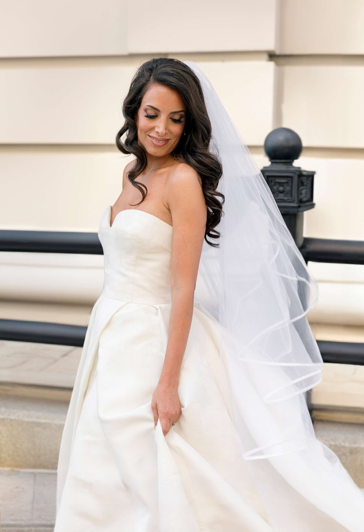 Bride in a strapless white wedding dress stands outdoors, holding her dress and smiling gently. Her long dark hair is styled in waves, and she wears a flowing veil. The background features a light-colored building and black railings.