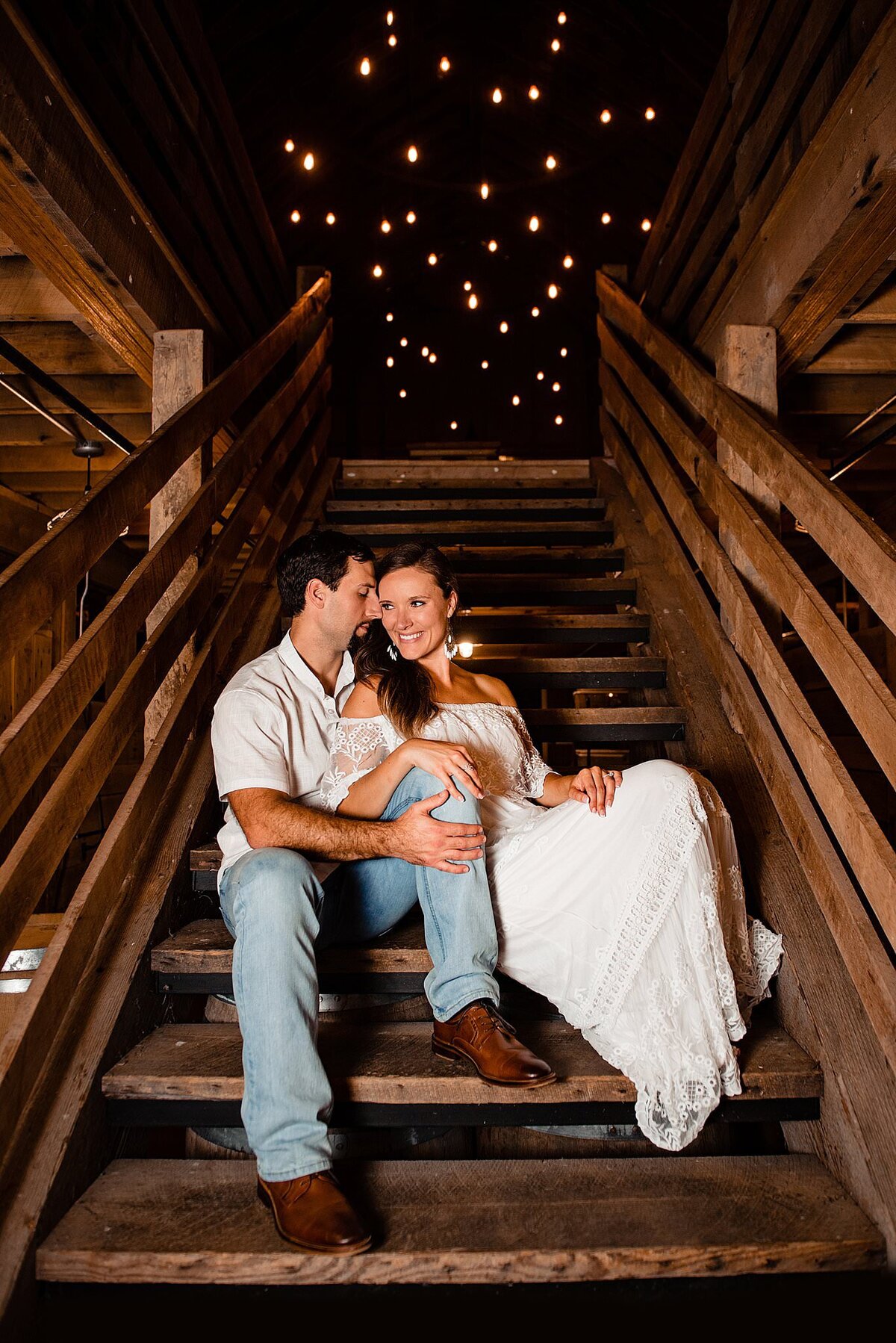 The bride and groom sit at the bottom of a wooden staircase. The groom, wearing brown shoes, light blue jeans and a white short sleeved golf shirt, whispers into the bride's ear. The bride leans against him and laughs at what he is whispering to her.  The bride is wearing an off the shoulder lace boho maxi dress with a wide lace ruffle around her shoulders. The wooden railing for the stairs is on either side of them and the string lighting at the top of the stairs looks like stars in the night sky.