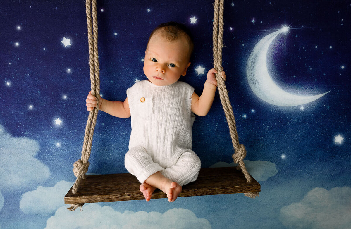 Newborn baby boy in a white sleeper on a swing with moon and stars in background