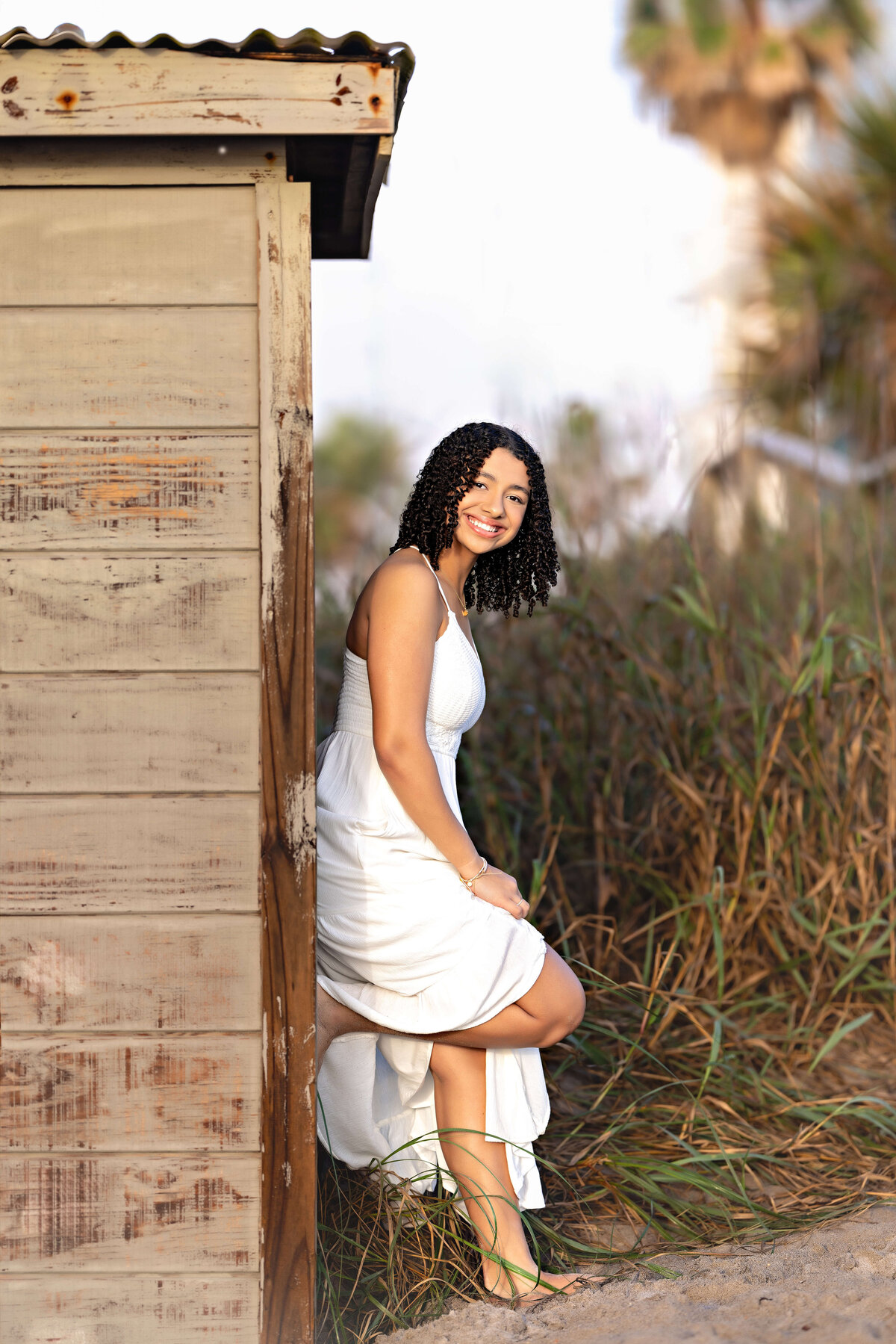 Orlando senior photographer capturing a high school girl smiling and leaning against a weathered beach shack during her Cocoa Beach sunrise session.