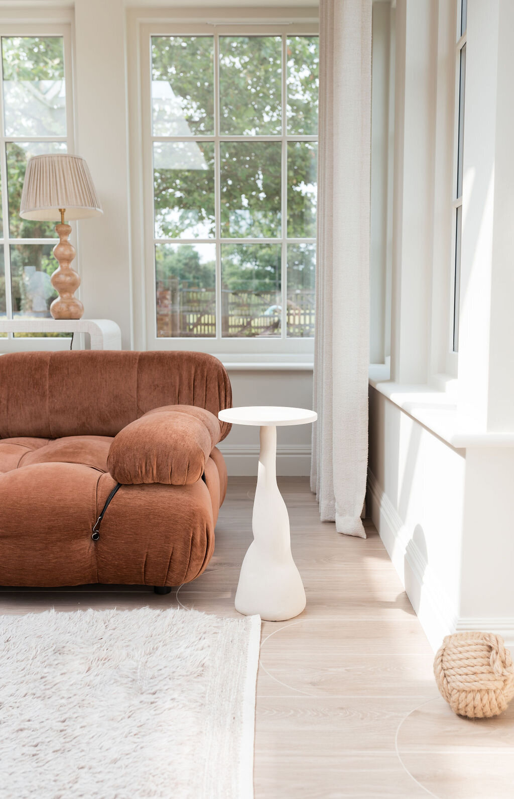 A sunlit living room featuring a rust orange, plush sofa, a sculptural white side table, and a decorative lamp on a wooden base. Large windows allow natural light to flood the room.