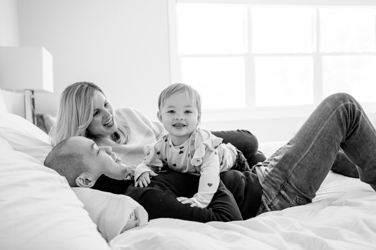 Family of Mom, Dad, and toddler snuggling in bed, toddler looking directly at camera