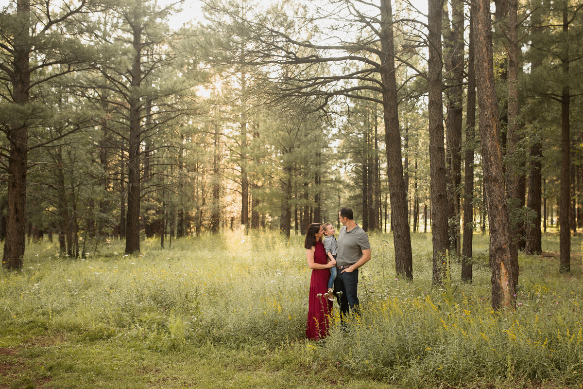 family of three standing in a grassy pine tree forest smiling at eachother