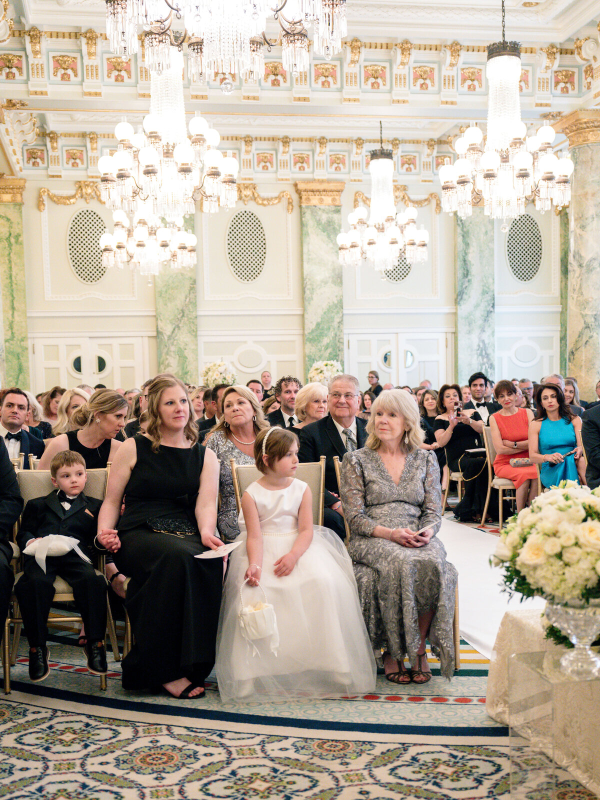 Guests sit in an elegant, ornate room with large chandeliers, attending a wedding ceremony. A woman in a gray dress and a young girl in a white dress are seated in the front row, along with other guests dressed formally.