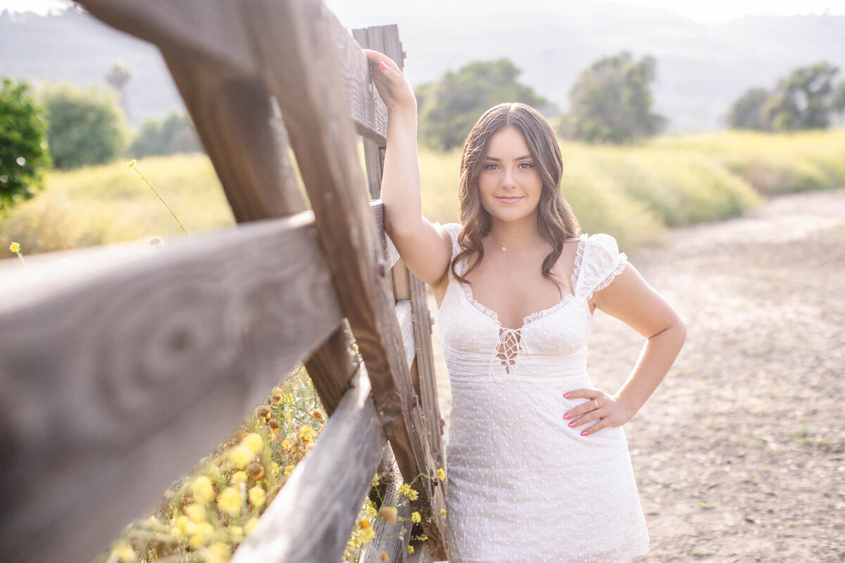 A woman in a white dress leans against a wooden fence in a sunny, rural landscape. She stands on a dirt path with fields and trees in the background, smiling softly at the camera.