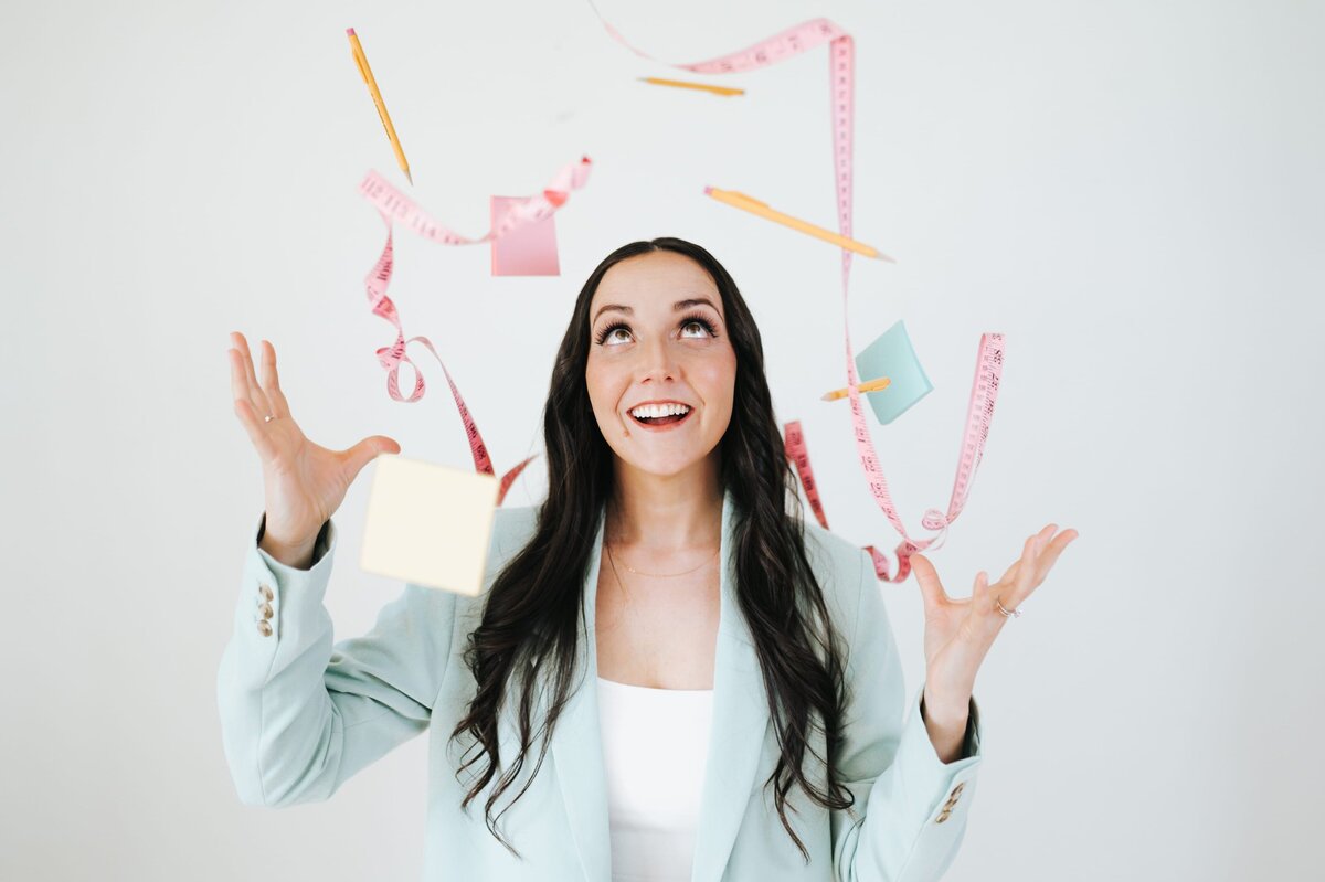 Woman in pastel blue blazer smiles up while reaching for pencil as several other pencils, post its and measuring tapes are suspended above her.