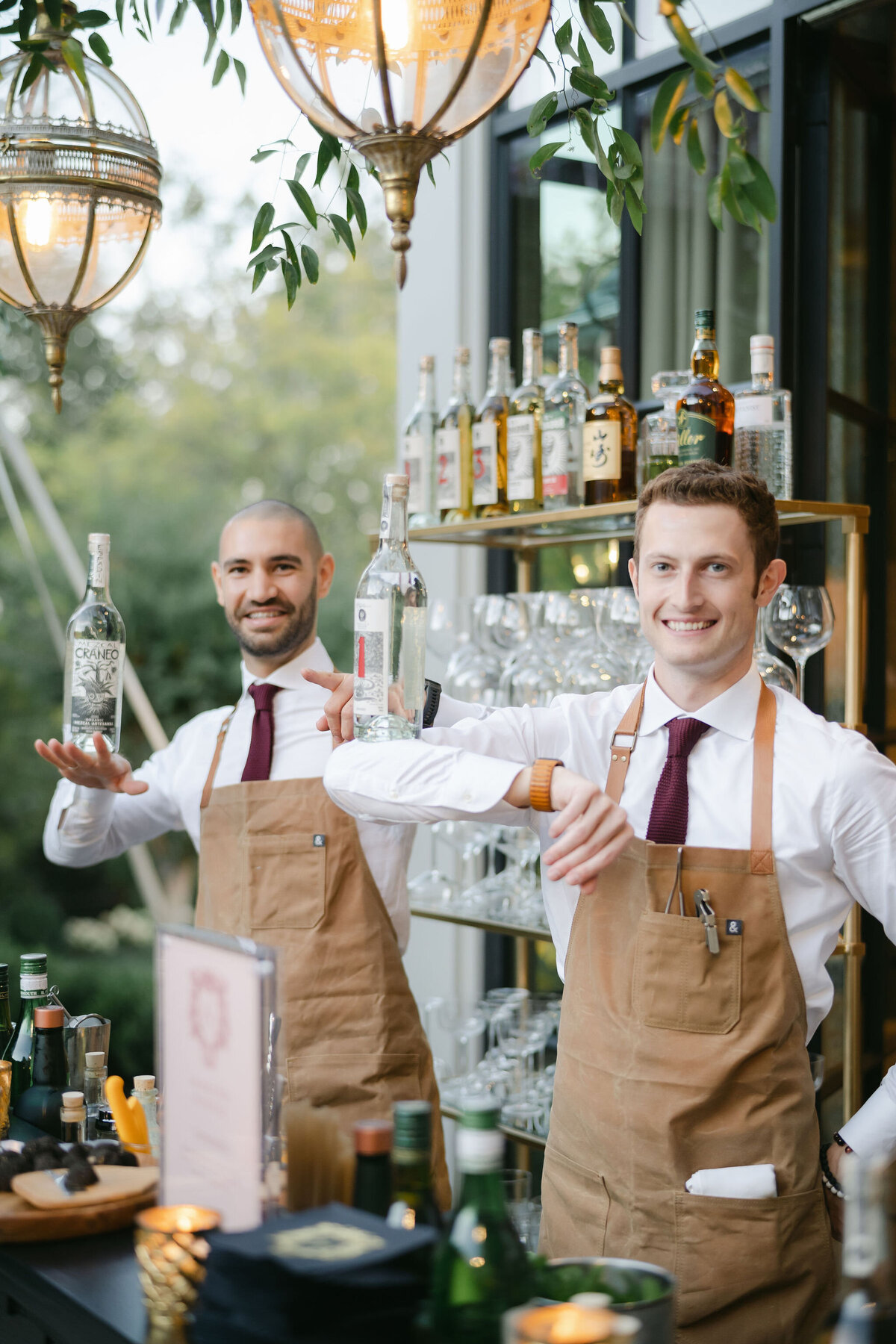 The photo was taken at a beautiful tented wedding in Virginia and captured by the talented photographer Elisabetta Marzetti. Clayton Films were present on-site as Videographers, capturing the special moments of the event. In the scene, two bartenders in crisp white shirts, red ties, and brown aprons stand behind a well-stocked bar, elegantly holding bottles of alcohol. The bar is meticulously arranged with a variety of bottles and glassware, creating an inviting atmosphere for guests. The space is adorned with hanging lights and lush greenery, complemented by the backdrop of large windows offering a view of the surrounding greenery, adding to the overall charm of the setting.