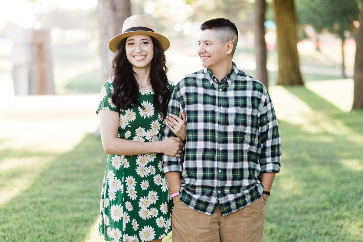 A lesbian couple walking closely together down a sidewalk and smiling during their engagement session in DFW, Texas. The woman on the left is wearing a green sundress decorated with daises and a straw hat. The woman on the right is wearing a green checkered shirt and khaki pants.