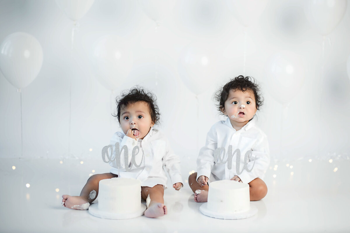 Twin one year old babies wearing white long sleeve shirts on white backdrop with white balloons eating a white iced cake