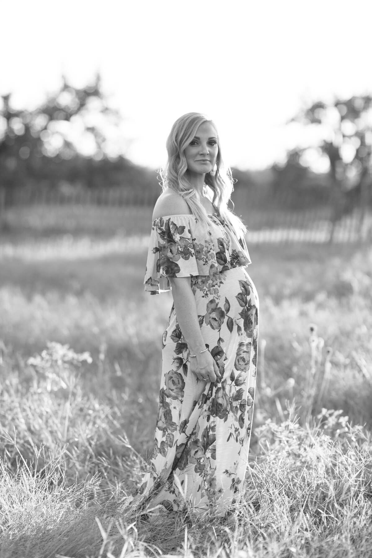 Pregnant woman looks into camera walking through a field in a floral dress.