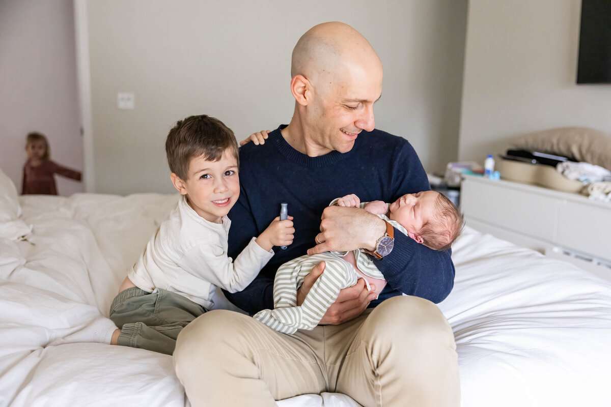 Dad holding baby boy with toddler at his side. Toddler daughter is standing in doorway