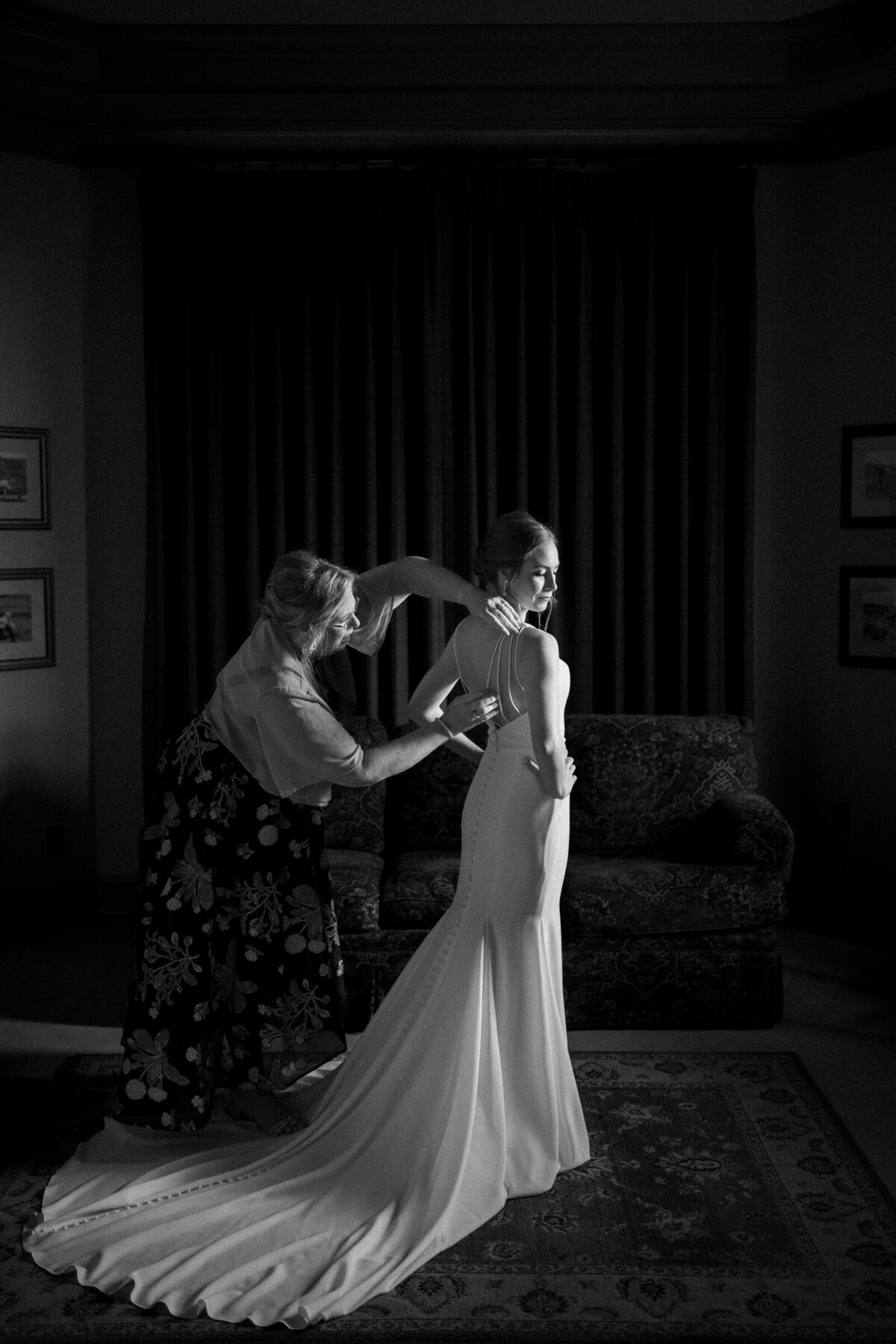 An elegant black and white photo captures a bride being assisted with her dress by a bridesmaid