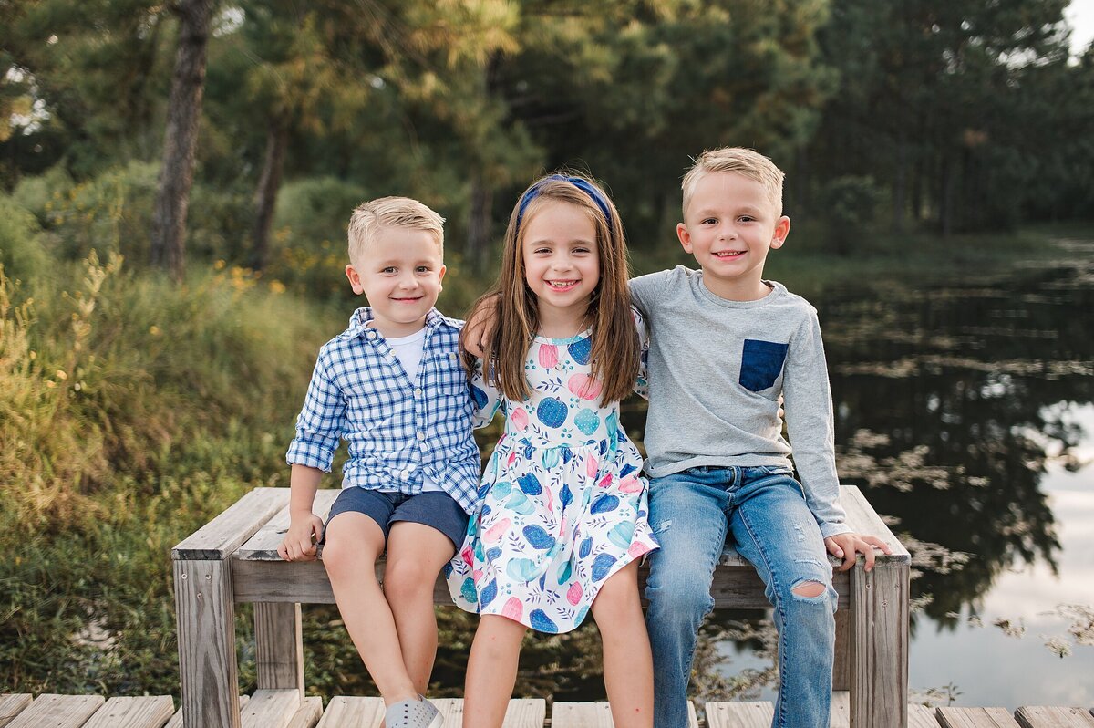 two brothers and their sister sitting on wooden bench by the water by Cypress Texas Photographer
