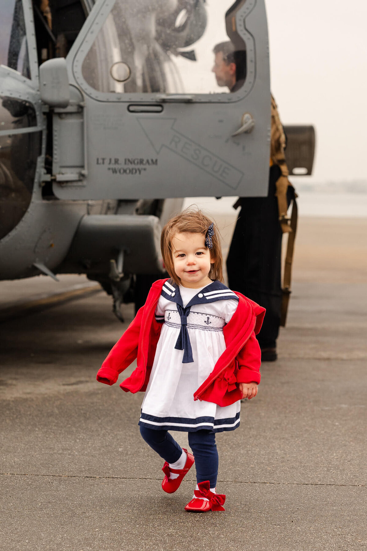 A toddler in a blue and white sailor dress and red jacket stands near a Navy helicopter.