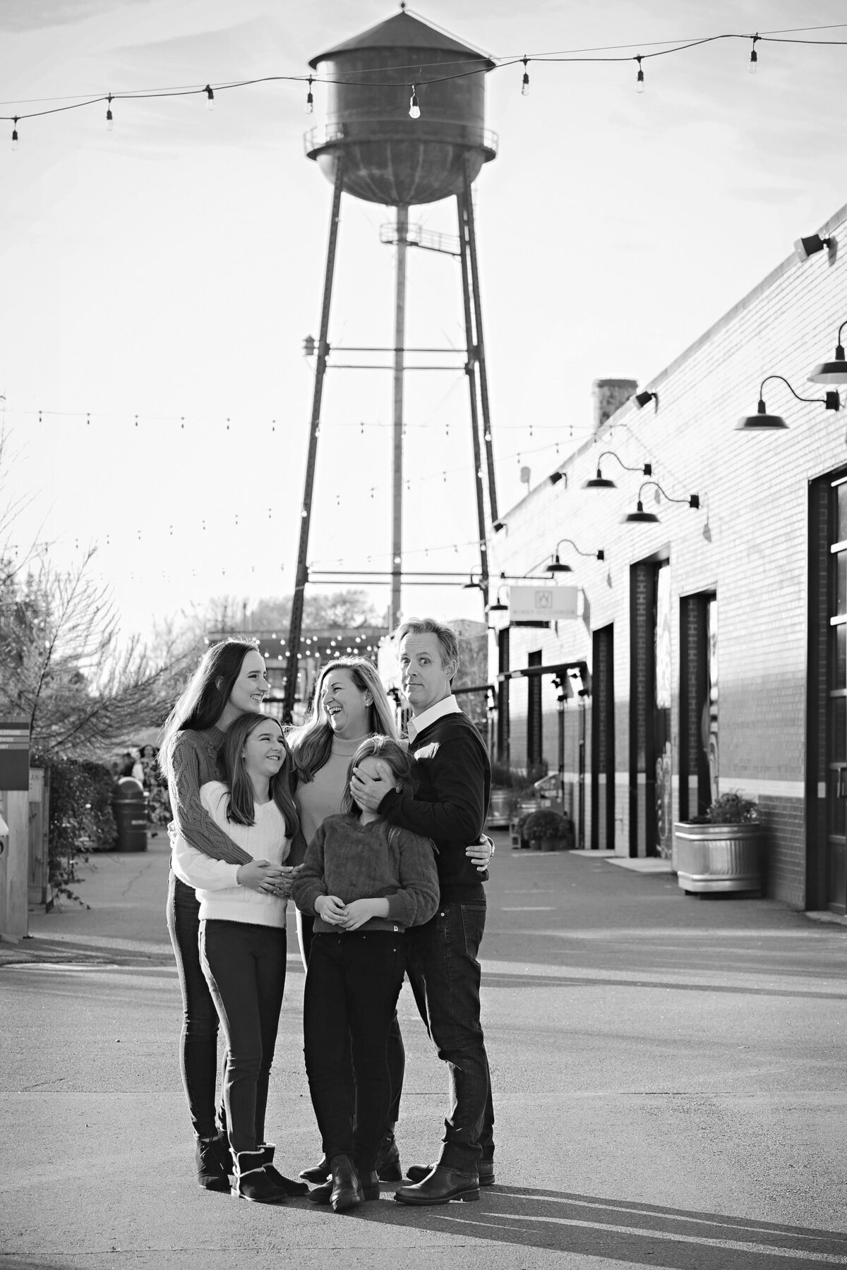 Black and white family portrait in front of industrial buildings and water tower at Camp North End Charlotte NC, everyone laughing as dad playfully covers youngest daughter’s face