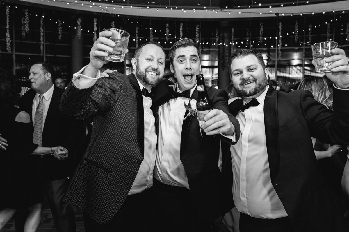 Three men cheering and holding drinks at a wedding reception